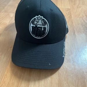 Svart Rögle keps från Bauer - Snygg svart keps med mesh på baksidan och broderad Rögle-logga framtill. Justerbar snapback för perfekt passform. Märket Bauer är broderat i vitt på sidan och bak. Perfekt för dig som gillar hockey och vill visa ditt stöd för Rögle.