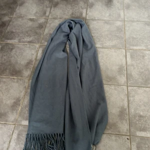 Mörkgrå halsduk med fransar - Säljer en stor och mysig mörkgrå halsduk med långa fransar i kanterna. Halsduken är enfärgad och har en klassisk rektangulär form som går att svepa flera varv runt halsen. Perfekt för kyliga dagar och enkel att matcha med olika outfits.