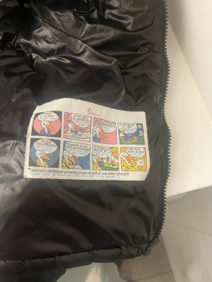 Svart pufferjacka från Moncler - Säljer en svart pufferjacka från Moncler med glansig finish och klassisk Moncler-logga på ärmen. Jackan har flera dragkedjeförsedda fickor och quiltad design. Perfekt för kalla dagar och riktigt snygg till streetwear-stilen.