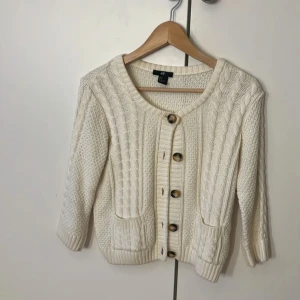 Vit stickad kofta från H&M, S - Superfin vit stickad kofta från H&M i storlek S. Koftan har snygga kabelstickade detaljer framtill, ribbade muddar och stora knappar. Den har två små fickor fram. Perfekt att slänga över en topp för en chill look.