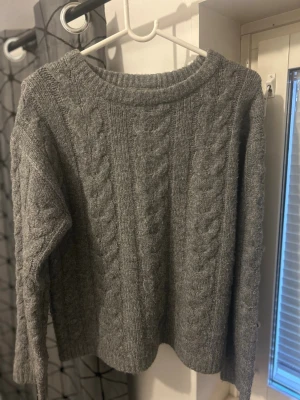 Grå kabelstickad tröja - Mysig grå stickad tröja med klassiskt kabelstickat mönster och rund hals. Tröjan har långa ärmar och ribbade muddar vid halsen. Perfekt för kyliga dagar och enkel att matcha med jeans eller kjol.