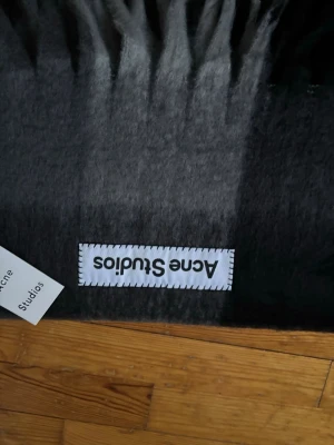 Mörkgrå halsduk från Acne Studios - Snygg och fluffig mörkgrå halsduk från Acne Studios med klassisk logotyp-patch och fransar i kanterna. Tillverkad i mjukt material som ger en lyxig känsla och passar perfekt till alla ytterkläder. En riktig statement-accessoar för dig som gillar stilrena detaljer.