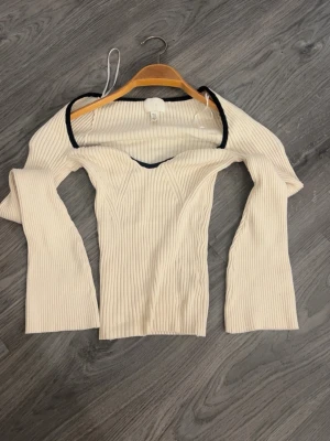 Ribbad offshoulder topp i beige/vit - Trendig ribbad offshoulder topp från H&M i ljusbeige med svarta kontrastkanter. Toppen har lång ärm och en snygg v-ringad skärning framtill. Perfekt för dig som gillar en stilren men ändå unik look.