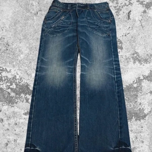 Blå jeans från MOD - Säljer ett par snygga blå jeans från MOD i storlek 29/32. De har en cool bootcut-stil med en lätt slitningseffekt på framsidan och baksidan. Jeansen har dragkedja och knappar framtill samt dekorativa sömmar som ger dem en unik look. Perfekta för en avslappnad vardagsstil!
