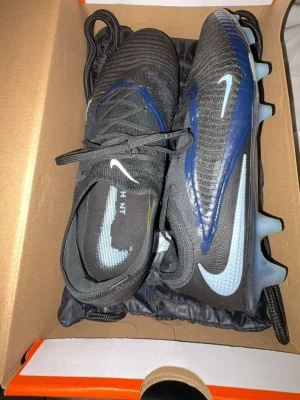 Nike Phantom Vision fotbollsskor svart/blå har använt 2 gånger typ köpte för 3000 - Säljer ett par Nike Phantom Vision fotbollsskor i svart med blå och vita detaljer. Skorna har snörning, syntetmaterial och en platt sula med dobbar för bra grepp på planen. Perfekta för dig som vill ha kontroll och stil på fotbollsplanen.
