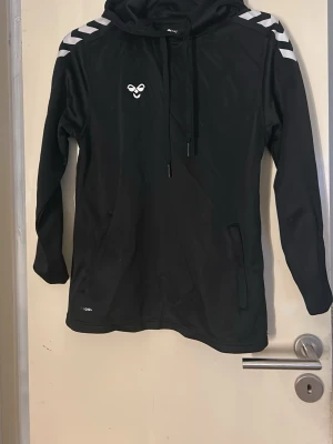 Svart hoodie från Hummel - Snygg svart hoodie från Hummel med vita detaljer på axlarna och logga på bröstet. Tröjan har huva med dragsko och praktiska fickor framtill. Tillverkad i ett sportigt, mjukt material som passar perfekt till chill eller träning. (Se bild 2) märket sitter löst och texten ”hummel” under har försvunnit. Annars i mycket fint skick