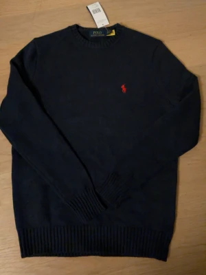 Mörkblå stickad tröja Polo Ralph Lauren S - Klassisk mörkblå stickad tröja från Polo Ralph Lauren med rund halsringning och ribbade muddar. Tröjan har den ikoniska röda logotypen broderad på bröstet. Perfekt för dig som gillar stilrena och tidlösa plagg. Aldrig använd storlek S
