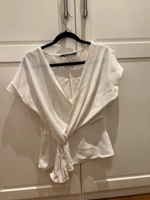 Vit omlottopp från Zara - Säljer en vit omlottopp från Zara med korta ärmar och knyt framtill. Toppen har en djup v-ringning och är gjord i ett mjukt, lätt material som ger ett snyggt fall. Perfekt att styla med jeans eller kjol för en fräsch look.