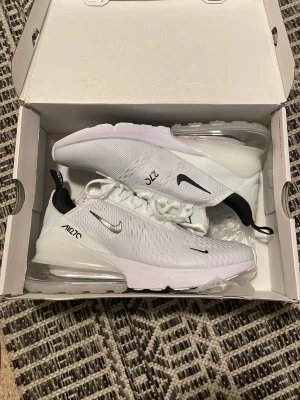 Nike Air Max 270 vita sneakers - Snygga vita Nike Air Max 270 sneakers med svart detalj på hälen och silverfärgad Swoosh. Skorna har en luftig mesh-överdel, synlig Air-enhet i sulan och klassisk snörning. Perfekta för dig som gillar stilrena och sportiga sneakers.