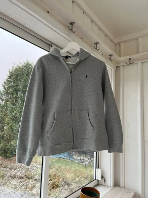 Grå zip hoodie från Polo Ralph Lauren - 18-20 - Snygg grå hoodie med dragkedja från Polo Ralph Lauren. Klassisk design med huva, två stora fickor framtill och den ikoniska mörkblå loggan broderad på bröstet. Tillverkad i mjukt sweatshirtmaterial som är perfekt för chill dagar. Passar runt 170