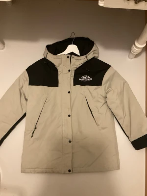 Beige och svart dunjacka  - Snygg beige och svart dunjacka med huva och dragkedja samt knappar framtill. Jackan har två snedställda fickor med dragkedja och logga på bröstet. Perfekt för kalla dagar och har en sportig look.