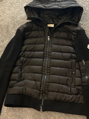 Svart Cardigan från Moncler med huva - Svart dunjacka från Moncler med quiltad framsida och huva. Jackan har långa stickade ärmar, dragkedja framtill och sidofickor med dragkedja. Perfekt för kalla dagar när du vill ha både stil och värme.
