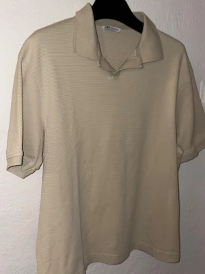 Beige pikétröja från Zara - Klassisk beige pikétröja från Zara med krage och korta ärmar. Tillverkad i ett mjukt, ribbat material som ger en avslappnad men ändå stilren look. Perfekt till jeans eller kjol för en enkel och snygg outfit.