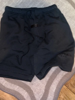Svarta shorts från Essentials - Säljer ett par svarta shorts från Essentials med dragsko i midjan och diskret logga på benet. Sköna och stilrena, perfekta för chill eller träning. Tillverkade i mjukt material för extra komfort.