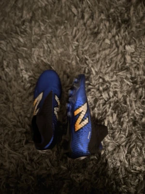 New Balance fotbollsskor elite - Säljer ett par blå fotbollsskor från New Balance med stora silvriga och gula detaljer på sidorna. Skorna har strumpliknande skaft och är tillverkade i syntetmaterial för bra passform och grepp på planen. Perfekta för dig som vill sticka ut på matchen! De är 39 1/2 men de är små i storlek så funkar för 39 och 39 1/2