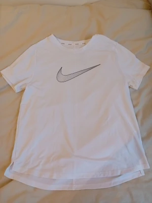 Vit Nike t-shirt med logga - Säljer en klassisk vit t-shirt från Nike med den ikoniska svarta swoosh-loggan på bröstet. T-shirten har rund halsringning och korta ärmar. Tillverkad i mjuk bomull för skön känsla och enkel stil.
