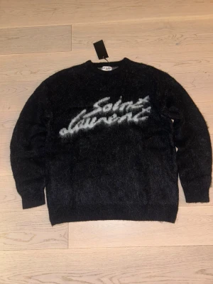 Svart Saint Laurent stickad tröja - Snygg svart stickad tröja från Saint Laurent med fluffig textlogga i vitt över bröstet. Tröjan har rund halsringning och långa ärmar. Materialet ser ut att vara mjukt och lite luddigt, perfekt för en trendig och avslappnad look. Den är oversized! Skulle pass någon som bär L bäst.