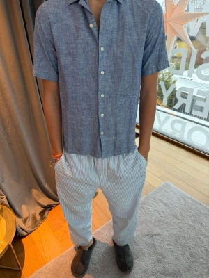 Blå linneskjorta med kort ärm - Snygg blå skjorta i linne med korta ärmar och klassisk krage. Skjortan har vita knappar framtill och en avslappnad passform. Perfekt för varma dagar och enkel att styla med olika outfits.