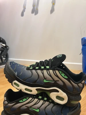 Nike Air Max Plus blå och gröna - Nike Air Max Plus sneakers i blått och svart med gröna detaljer och svart mesh. Snygg vågig design på ovandelen, gröna swoosh-loggor och synliga Air Max-enheter i sulan. Skorna har snörning och en sportig vibe som sticker ut.