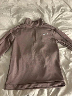 Ljuslila träningströja från Nike - Snygg ljusbrun långärmad träningströja från Nike med halv dragkedja och hög krage. Perfekt för löpning eller gym. Tröjan har en stilren design med Nike-logga på bröstet och är tillverkad i ett mjukt, stretchigt material.