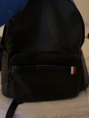 Svart ryggsäck från Tommy Hilfiger - Snygg svart ryggsäck från Tommy Hilfiger i skinnimitation med klassiska Hilfiger-detaljer i rött, vitt och blått. Rymligt huvudfack, framficka med dragkedja och justerbara axelband. Perfekt för skolan eller stan. Får plats med MacBook och i mycket bra skick!