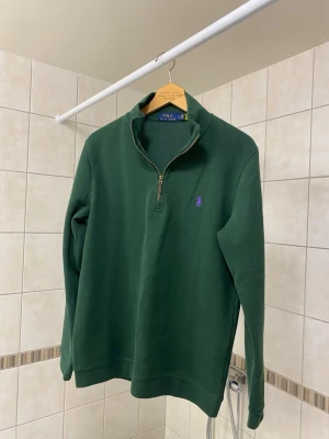 Polo Ralph Lauren Half Zip - Mörkgrön half-zip tröja från Polo Ralph Lauren med lila broderad logga på bröstet. Tröjan har hög krage, dragkedja framtill och är tillverkad i mjukt bomullsmaterial. Perfekt för en avslappnad men ändå stilren look. Helt ny i princip.