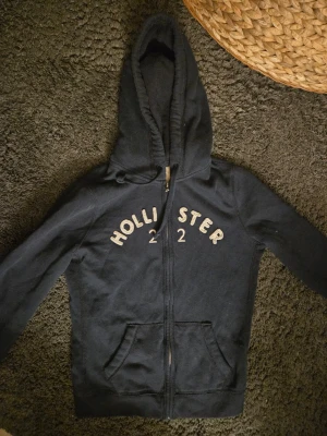 Mörkblå hoodie från Hollister - Mörkblå hoodie från Hollister med dragkedja framtill och stor huva. Broderad text 'HOLLISTER 22' i vitt över bröstet. Två fickor framtill och ribbade muddar vid ärmslut och nederkant. Perfekt för chill dagar.