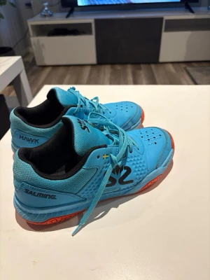 Blå Salming Hawk inomhusskor - Säljer ett par blå Salming Hawk inomhusskor med svart och orangea detaljer. Skorna har snörning, ventilerande meshmaterial och stabil sula som passar perfekt för innebandy eller andra inomhussporter. Snygg design med svart logga på sidan.