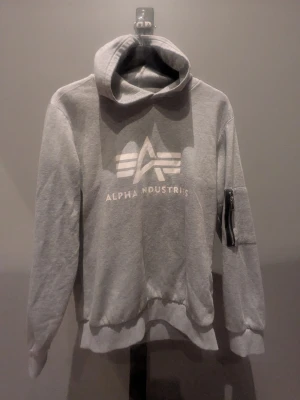 Grå hoodie Alpha Industries - Snygg grå hoodie från Alpha Industries med stor logga framtill och dragkedjeficka på ena ärmen. Klassisk huva och ribbade muddar vid ärmslut och nederkant. Perfekt för en avslappnad och cool stil.