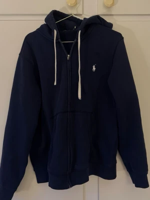 Marinblå hoodie från Polo Ralph Lauren - Snygg marinblå hoodie från Polo Ralph Lauren med dragkedja, vit broderad logga på bröstet och vita snören i huvan. Tillverkad i mjukt bomullsmaterial och har två fickor framtill. Perfekt för en avslappnad och stilren look.