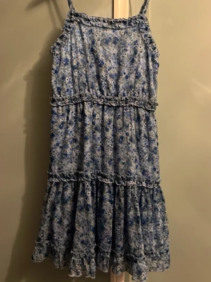 Blommig blå kortklänning med volang - Söt kortklänning med blått och vitt blommönster från Lindex, tunna axelband som går att justera och volangdetaljer. Aldrig använd, har lappen kvar.