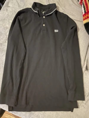 Svart långärmad polotopp från Lee - Klassisk svart långärmad polotopp från Lee med vit kontrastkant på kragen och knappslå framtill. Regular fit och diskret Lee-logga broderad på bröstet. Perfekt till jeans eller chinos för en clean look.