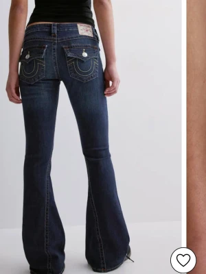Mörkblå bootcut jeans från True Religion - Snygga mörkblå bootcut jeans från True Religion med ikoniska kontrastsömmar och stora fickor bak med lock och knappar. Jeansen har låg midja och klassisk storleken är w24 