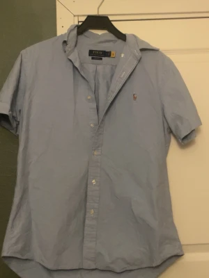 Blå kortärmad skjorta från Polo Ralph Lauren - Snygg ljusblå skjorta från Polo Ralph Lauren med korta ärmar och klassisk krage. Skjortan har knappar framtill och den ikoniska broderade loggan på bröstet. Tillverkad i mjuk bomull som känns skön mot huden. Perfekt för en avslappnad men stilren look. Kom privat för fler bilder eller frågor.mvh