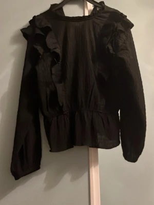 Svart blus med volanger - Snygg svart blus med volangdetaljer på axlarna och rynkad struktur i tyget. Blusen har rund halsringning och långa ärmar med resår vid ärmsluten. Perfekt för dig som gillar en trendig och stilren look. Har använt den några gånger men är i väldigt bra skick 
