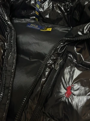 Svart pufferjacka från Polo Ralph Lauren - Snygg svart pufferjacka från Polo Ralph Lauren med glansig finish och huva. Jackan har dragkedja framtill, flera fickor med dragkedja och röd broderad logga på bröstet. Perfekt för kalla dagar och har en riktigt modern vibe.
