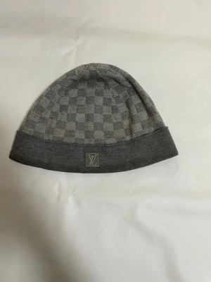 Grå Louis Vuitton mössa - Snygg grå mössa från Louis Vuitton med klassiskt rutmönster och vikt kant. Diskret LV-logga framtill och mjukt material som känns lyxigt. Perfekt accessoar för dig som gillar exklusiv streetstyle.