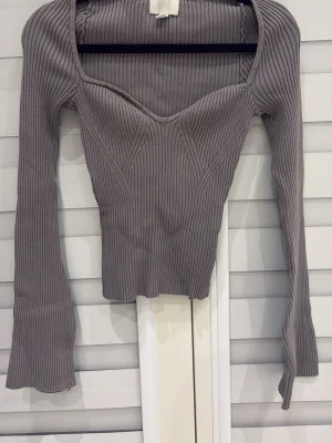 Ribbad grå croppad topp - Trendig ribbad topp i grått med croppad passform och långa ärmar. Toppen har en snygg sweetheart-urringning och markerade sömmar framtill som ger en cool siluett. Perfekt för att styla med höga jeans eller kjol.