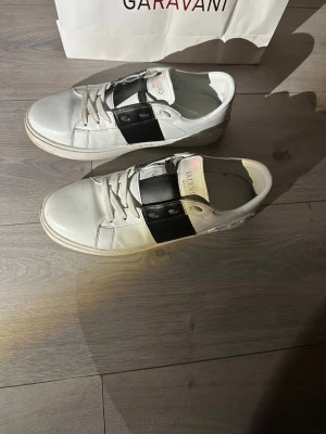 Valentino Garavani vita sneakers - Säljer ett par vita Valentino Garavani sneakers med svart band över mitten och coola nitar på hälen. Skorna har snörning, rund tå och platt sula. Tillverkade i skinn och har en stilren, modern look som passar till det mesta.