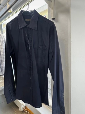 Mörkblå skjorta från Dressmann - Klassisk mörkblå skjorta från Dressmann i 100% bomull. Skjortan har lång ärm, bröstficka med knapp och knappar längs hela framsidan. Perfekt till jeans eller chinos för en clean look. Solmärken i kragen.
