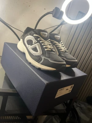 Svarta och vita Dior B30 skor - Säljer ett par svarta Dior B30 sneakers med vita och grå detaljer. Skorna har en chunky sula, meshpaneler och stora reflekterande Dior-loggor på sidorna. Klassisk snörning och sportig vibe. Perfekta för dig som vill ha något exklusivt och trendigt.