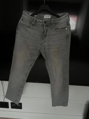 Grå jeans från Jack & Jones - Snygga grå jeans från Jack & Jones med klassisk femficksdesign och raka ben. Jeansen har en diskret tvättad look och är tillverkade i mjukt denimtyg som ger en skön känsla. Perfekta för en avslappnad och stilren outfit. Lite uppsydda 