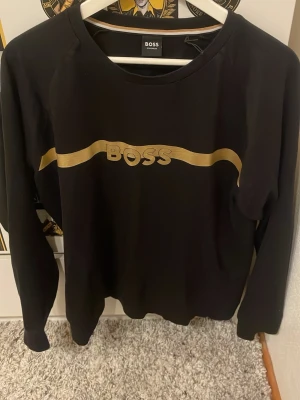 Svart sweatshirt från BOSS med guldtryck - Snygg svart sweatshirt från BOSS med ett guldigt vågformat tryck och logga över bröstet. Tröjan har rund halsringning och långa ärmar. Perfekt för dig som gillar stilrena plagg med en exklusiv touch.