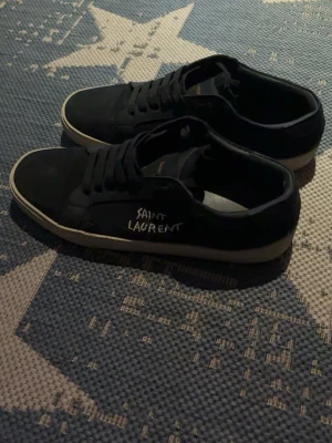 Svarta Saint Laurent sneakers - Säljer ett par svarta sneakers från Saint Laurent med vit logga på sidan och vit sula. Klassisk låg modell med snörning och rund tå. Perfekta för dig som gillar stilrena och tidlösa skor.