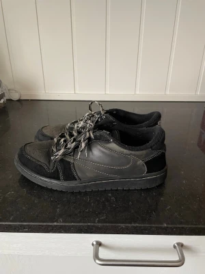 Travis Scott Low Black Phantom  - Säljer ett par svarta sneakers med vita kontrastsömmar och svartvita snören. Skorna har rund tå, perforerade detaljer på tån och platt sula. Materialmix av skinn och mocka ger en schysst look som passar till det mesta.