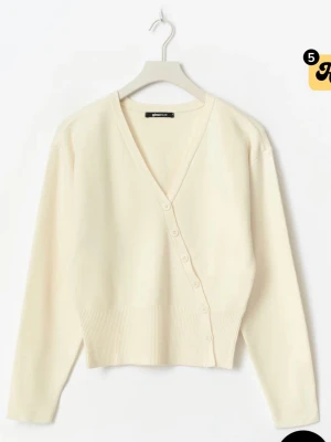 Krämvit kofta från Gina Tricot - Snygg krämvit kofta från Gina Tricot med v-ringning och knappar längs sidan framtill. Koftan har långa ärmar och bred ribbad mudd nedtill för en modern look. Perfekt att styla med jeans eller kjol för en clean och enkel outfit.