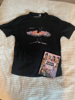 Ariana Grande T-shirt + bok - Det är en T -shirt och en bok om Ariana Grande.  Bild 4 är en bild på framsidan av tröjan och bild 5 är en bild på baksidan av tröjan. T-shirten är lite större än en Xs utan mer en S, och båda är i mycket bra skick.