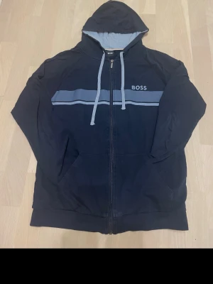 Mörkblå hoodie från BOSS med dragkedja - Snygg mörkblå hoodie från BOSS med dragkedja framtill, ljusblåa snören och huva. Bröstet pryds av en bred ljusblå rand och BOSS-loggan. Hoodien har fickor på sidorna och är tillverkad i mjukt material för en bekväm känsla.