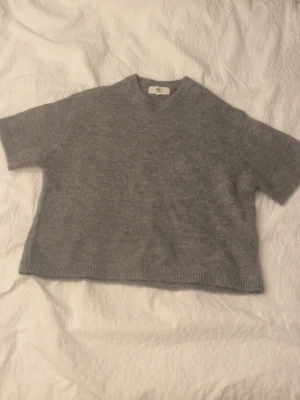 Grå stickad tröja Gina Tricot - Kort, grå stickad tröja från Gina Tricot i mjuk ullblandning. Tröjan har rund halsringning, korta ärmar och en avslappnad, boxig passform. Perfekt att styla med höga jeans eller kjol för en trendig look.