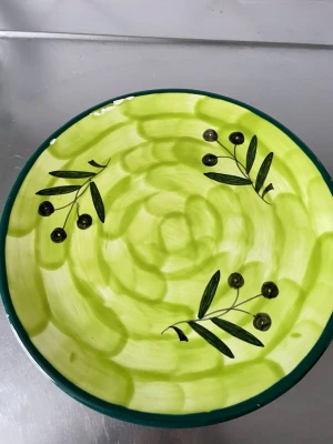 Grön keramikfat med olivmotiv - Unikt keramikfat i gröna nyanser med handmålat olivmotiv och mörkgrön kant. Fatet har rund form och kan användas både som dekoration eller serveringsfat. Baksidan har upphängningsanordning för vägglite fel i glaseringen enligt bild 